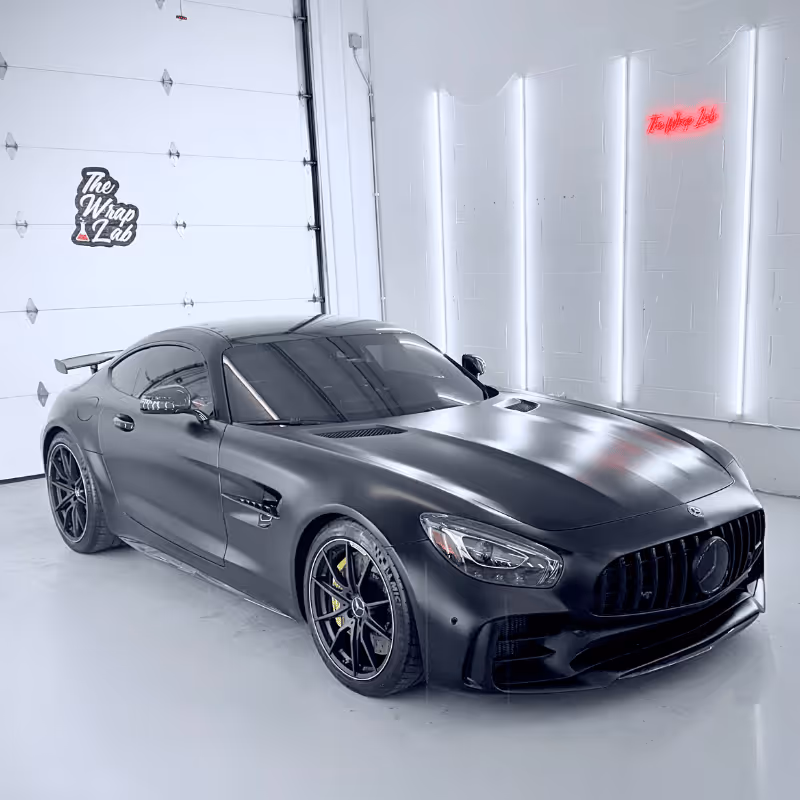 A sleek black Mercedes-Benz sports car sits in a well-lit garage space with white walls. The vehicle features a matte finish, prominent grille, and large alloy wheels. A neon sign reading 'The Wrap Lab' is visible in the background.