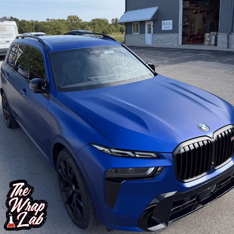 A blue matte-finish BMW SUV parked in front of a grey building. The vehicle's sleek design features bold lines, a distinctive kidney grille, and black accents. The Wrap Lab logo is visible in the corner of the image.