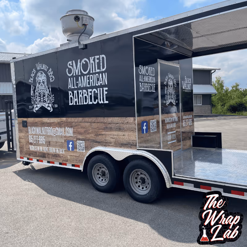 A black food trailer advertising "Smoked All-American Barbecue" with a logo featuring a skull and crossbones on the side. The window is open, revealing a spacious interior. The trailer is parked on a gravel surface under a clear blue sky.