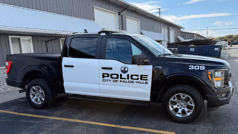 A black and white police pickup truck parked beside a building. The side displays "POLICE CITY OF PALOS HILLS" with a badge symbol on the door. The truck has four doors and rugged tires, set against a backdrop of a commercial building.