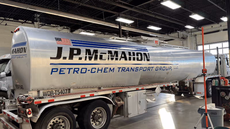 A shiny silver tanker trailer parked inside a garage, displaying the name 'J.P. McMahon' and 'PETRO-CHEM Transport Group, LLC' in bold blue lettering. The interior features overhead lights and various equipment, creating an industrial atmosphere.