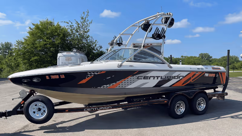 A sleek white and black Centurion boat with striking orange accents, mounted on a double-axle trailer. The boat features a wakeboard tower with multiple holders, parked on a paved surface with a background of lush greenery and a clear blue sky.