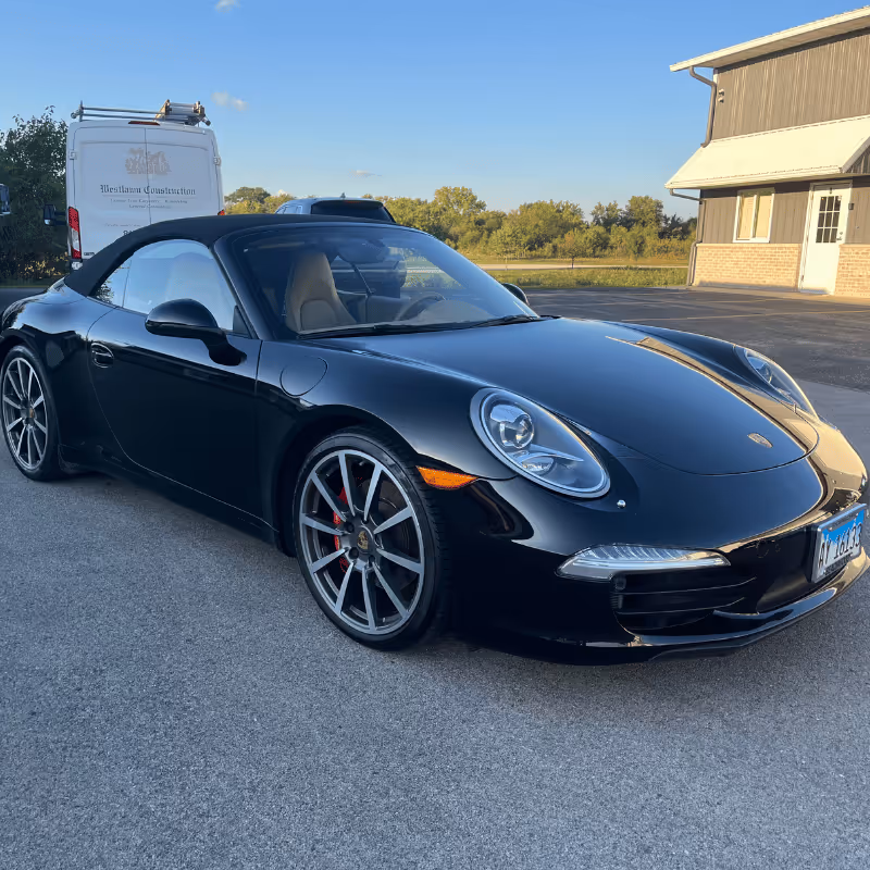 A sleek black convertible sports car parked on a paved surface, showcasing its shiny exterior and modern design. The vehicle features distinctive round headlights and sporty alloy wheels, set against a backdrop of trees and a building.