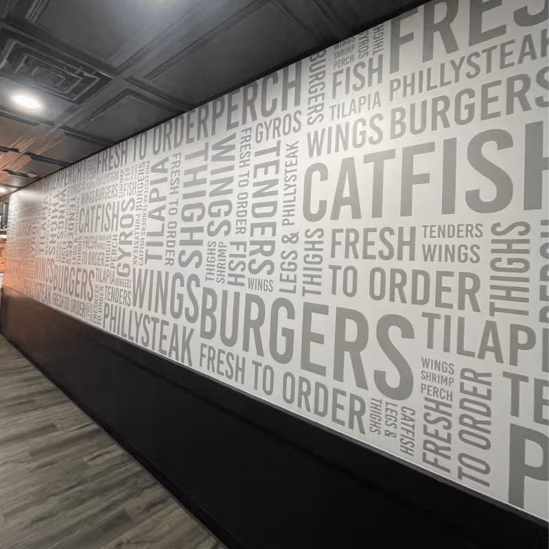 A wall covered with large text displaying food items such as 'Catfish', 'Wings', 'Burgers', and 'Philly Steak' in a modern restaurant setting, emphasizing freshness and made-to-order meals for diners.