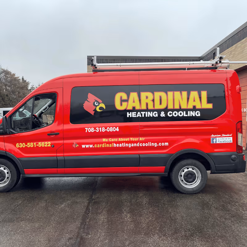 A red service van with the logo of Cardinal Heating & Cooling on its side. The van features contact numbers and a website, parked in a commercial area, showcasing its branding for HVAC services.