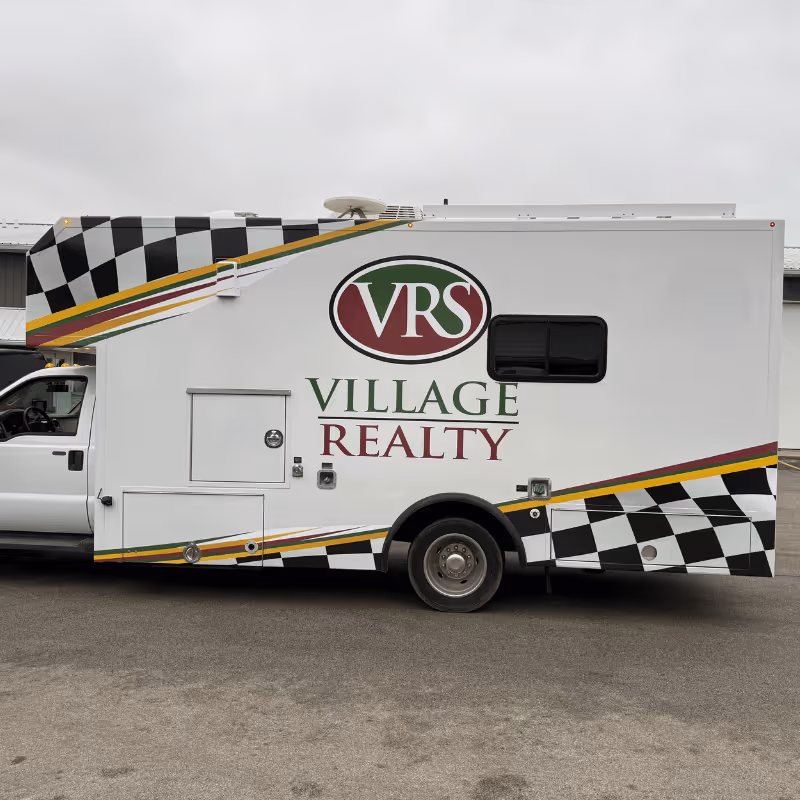 A side view of a white vehicle featuring the logo "VRS" and the text "Village Realty" in red and green. The vehicle has a checkered pattern in black and white along the bottom, set against a gray sky.