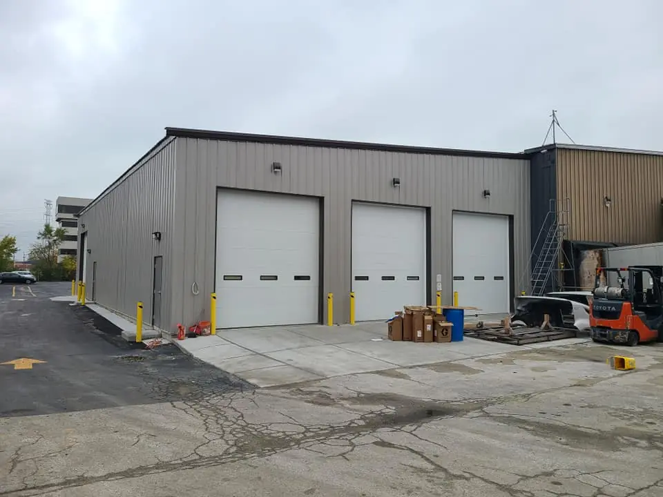 Exterior of the metal-clad warehouse addition with three overhead bay doors, yellow bollards, and an active loading area.