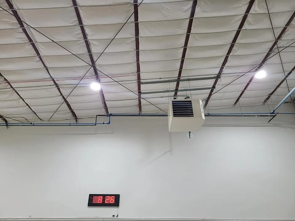 Ceiling-mounted unit heater with ductwork routed through the open steel roof framing of the warehouse addition.