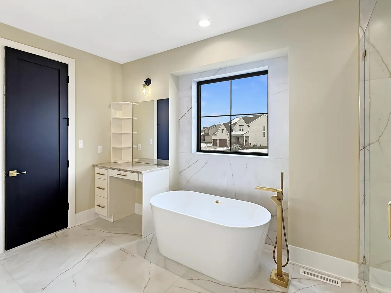 Freestanding soaking tub tucked beneath a marble-tiled window, next to a built-in makeup vanity.