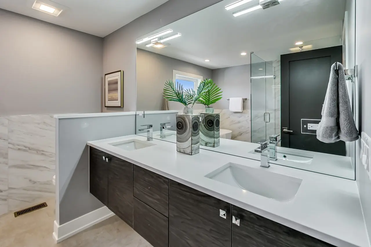 Double-sink bathroom vanity with a white quartz counter, dark wood cabinetry, and a glass shower beyond.