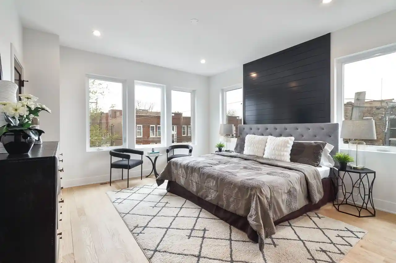 Primary bedroom with a tufted gray headboard, black shiplap accent wall, and rows of windows framing city rooftops.
