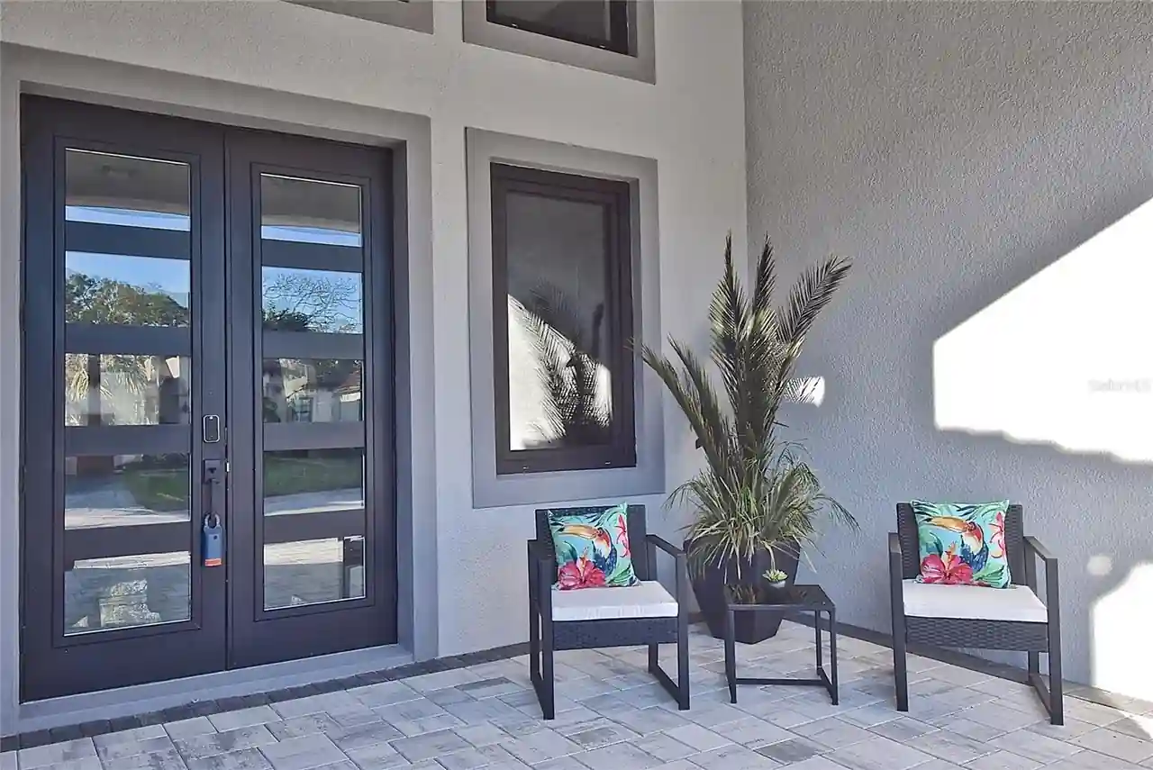 Covered entry patio with wicker lounge chairs, a potted palm, and glass double doors.