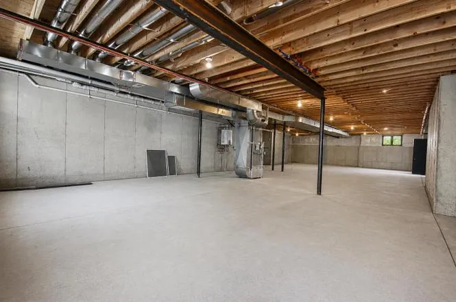 Wide-open unfinished basement with wood joists, exposed ductwork, and steel support posts.