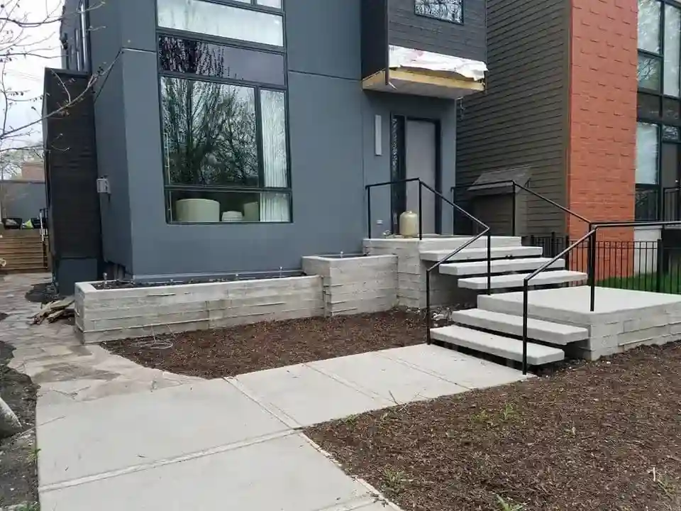 Front entrance of the addition with new concrete steps, black metal railing, and fresh landscaping beds