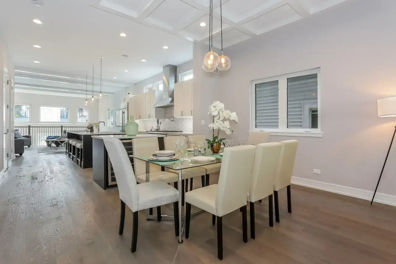 Dining area with glass table, upholstered chairs, pendant lighting, and open kitchen view