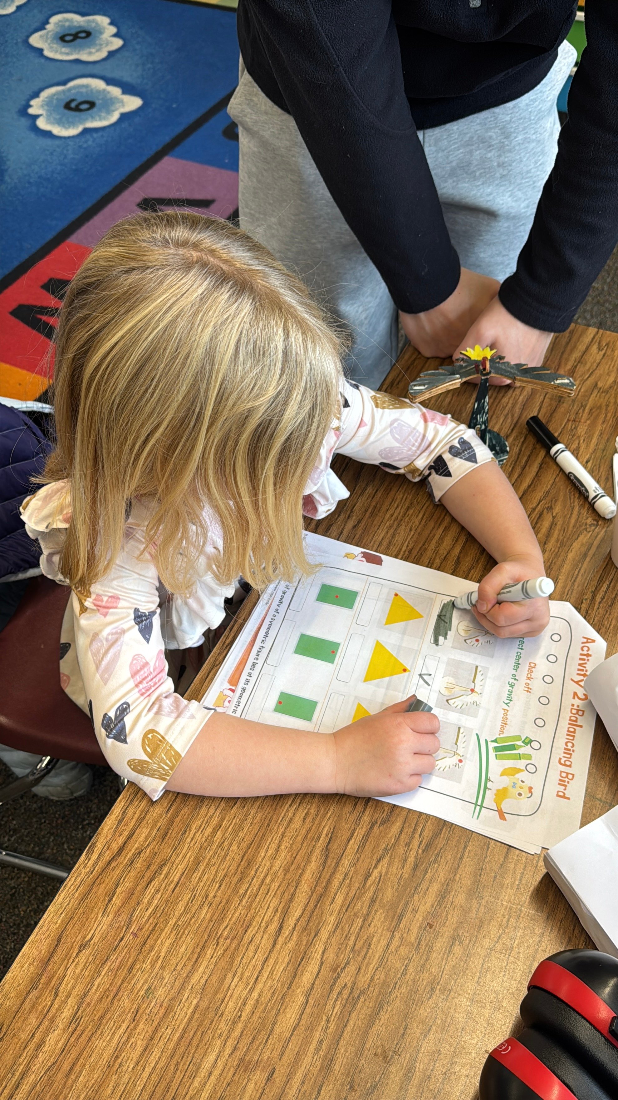 Child with blonde hair using a marker to complete a balancing bird activity worksheet on a wooden table.