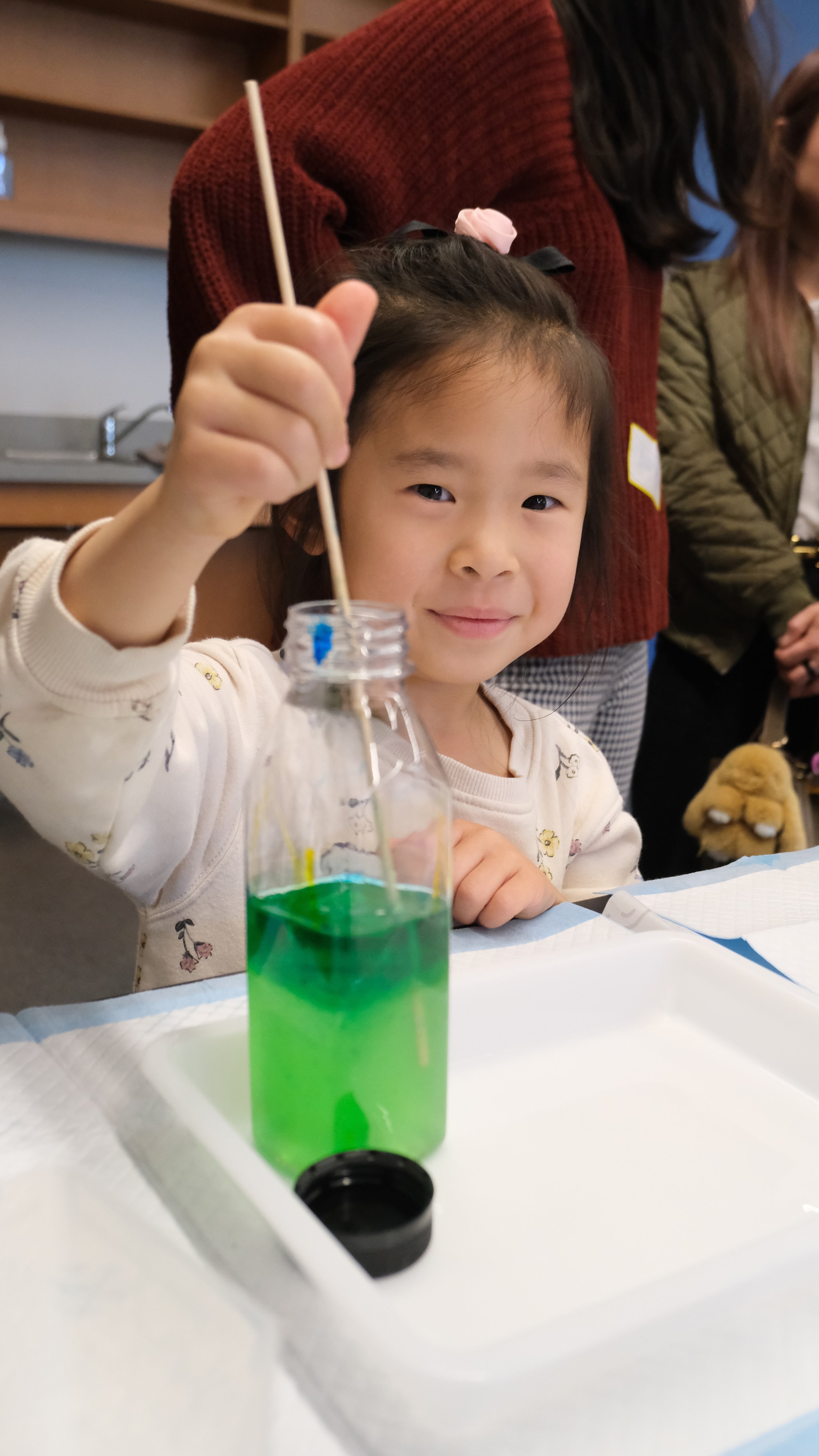Young girl stirring a green liquid inside a plastic bottle with a wooden stick at a table.