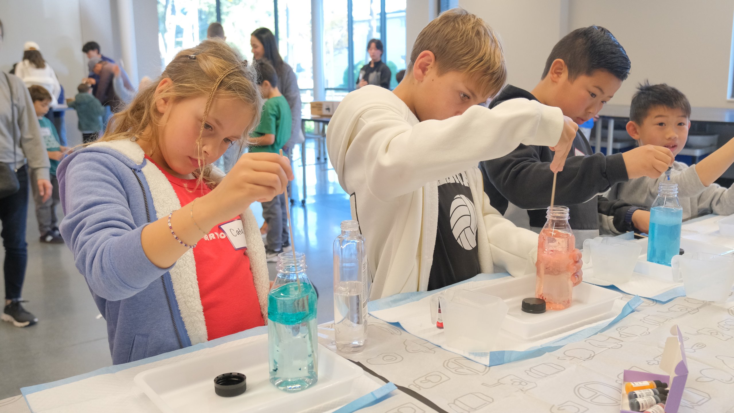 Four children engaged in a hands-on science activity mixing colored liquids in bottles at a classroom table.