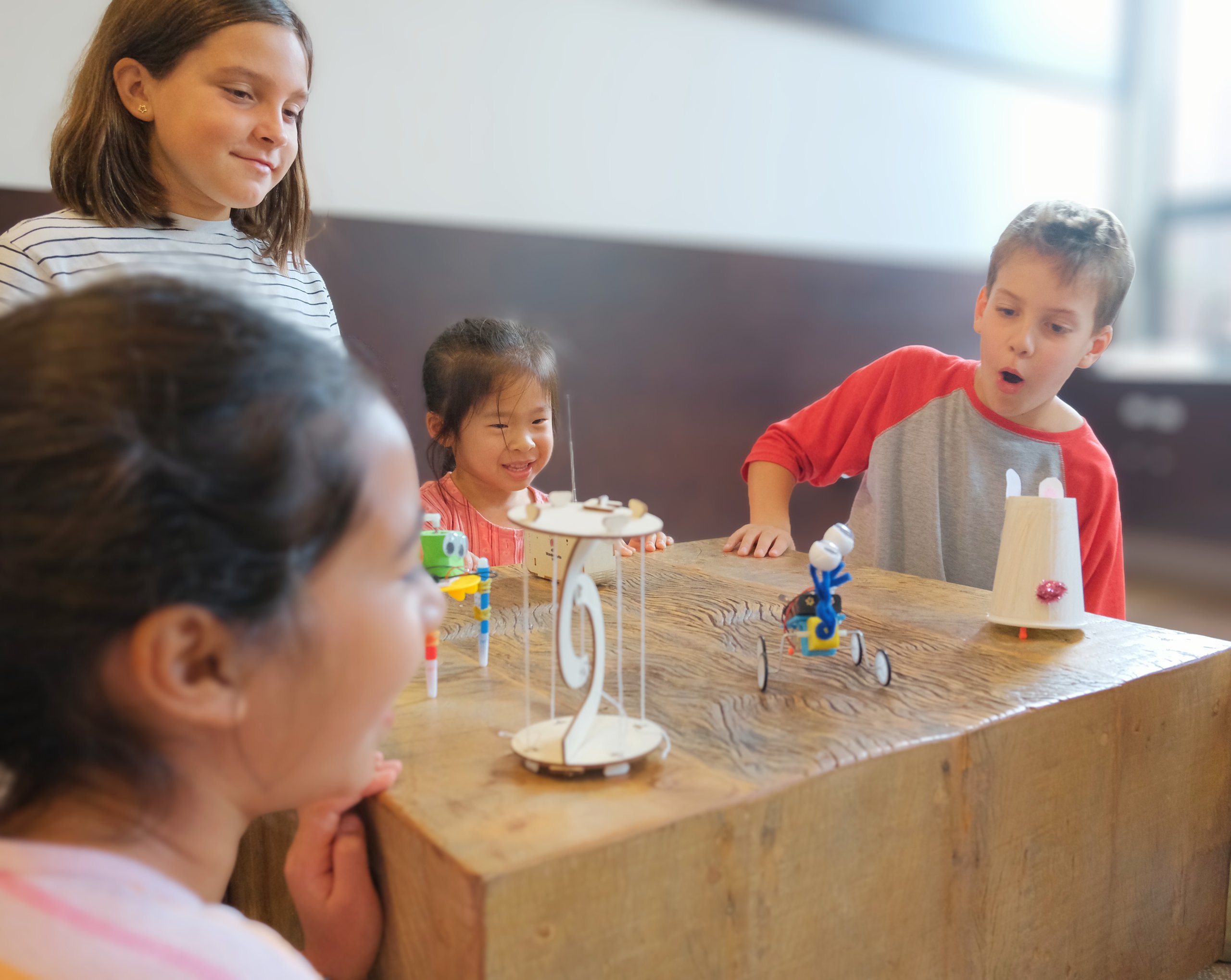 Four children gathered around a wooden table, excitedly looking at small robotic toys and crafts placed on the table.