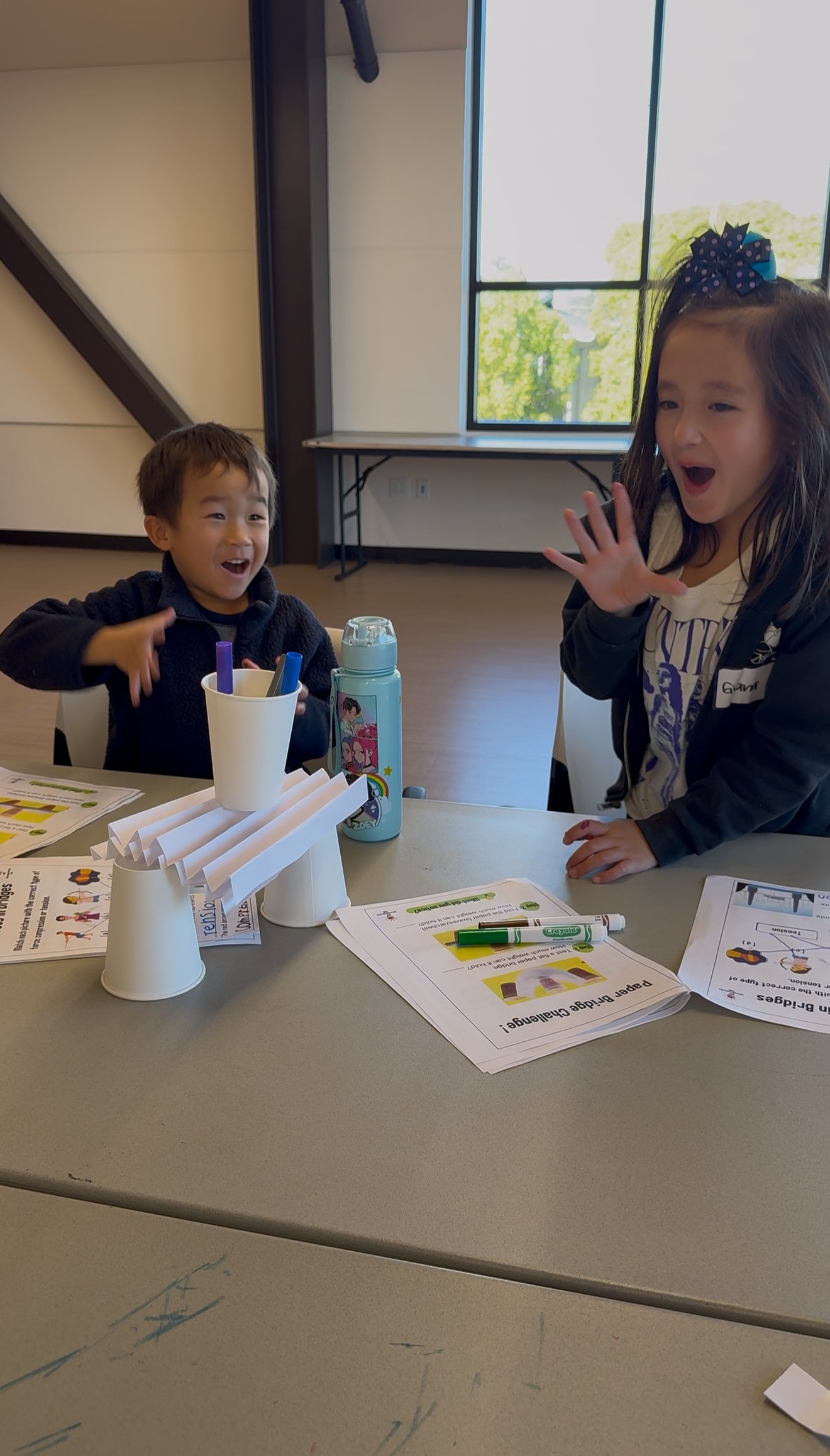 Two children reacting excitedly at a table with paper cup structures and coloring materials.