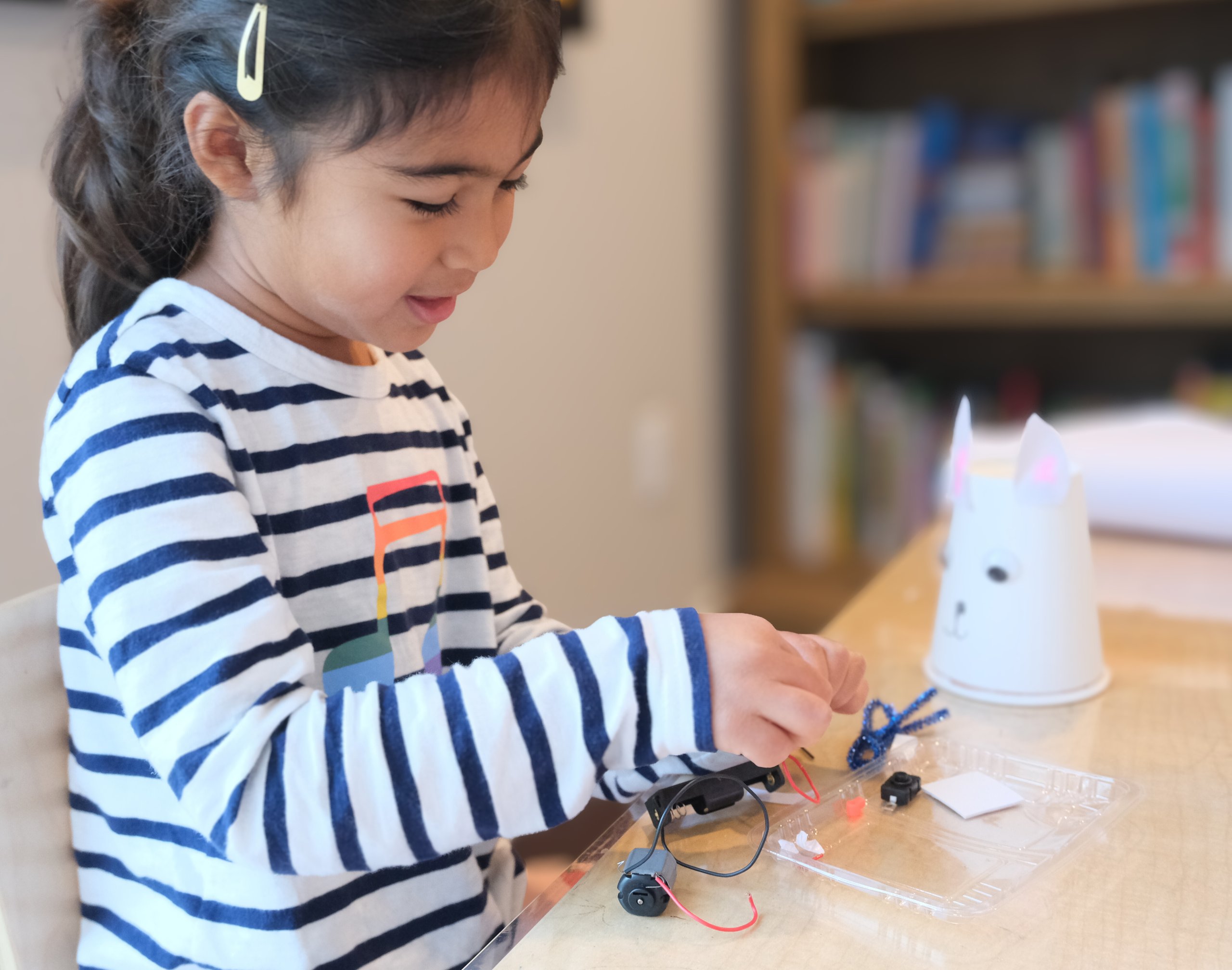 Young girl with a striped shirt assembling craft materials and wires at a table.
