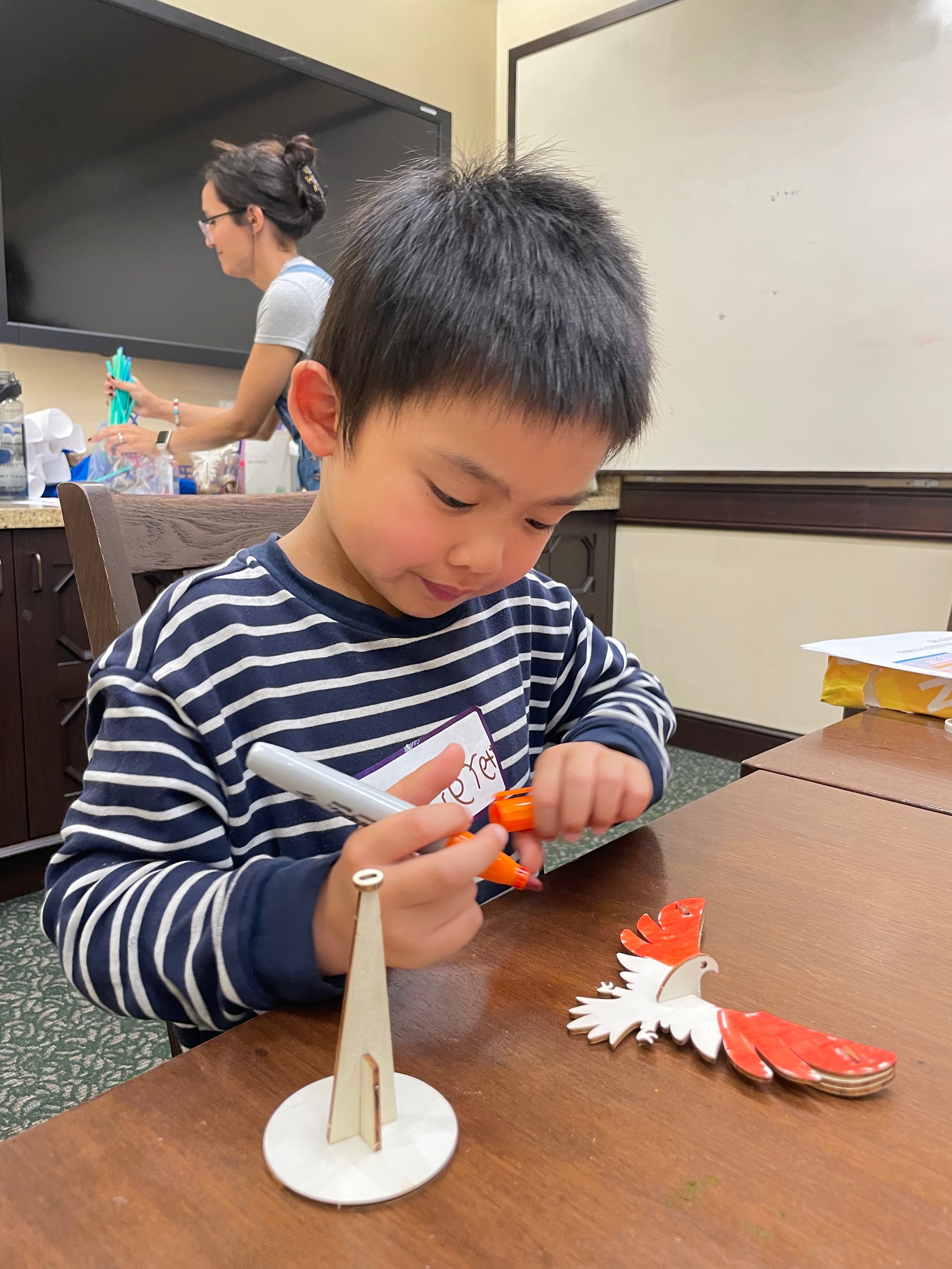 Young boy in a striped shirt coloring a wooden bird figure with orange markers at a table.