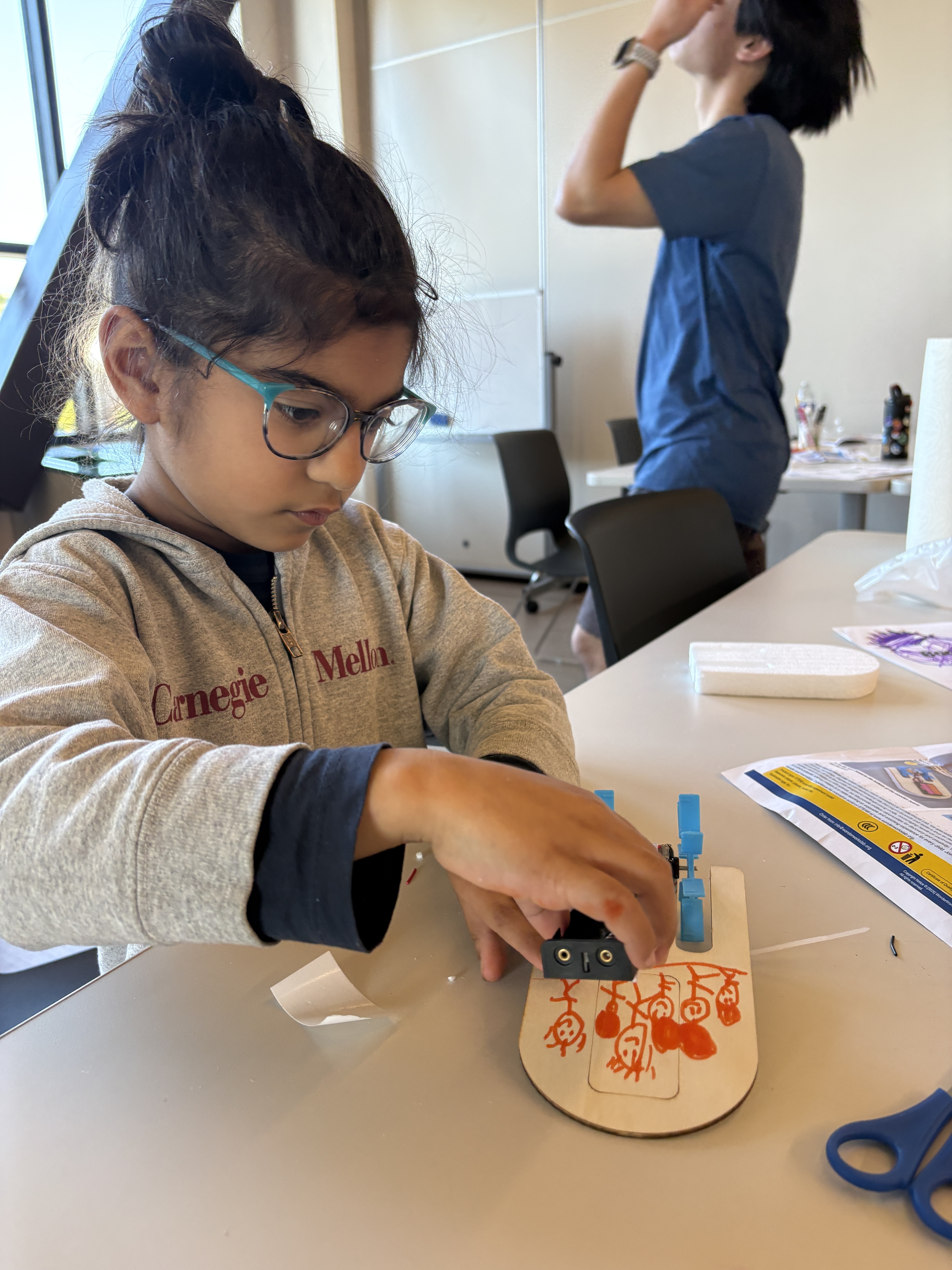 A child wearing glasses and a gray Carnegie Mellon hoodie assembling a small craft project on a table.