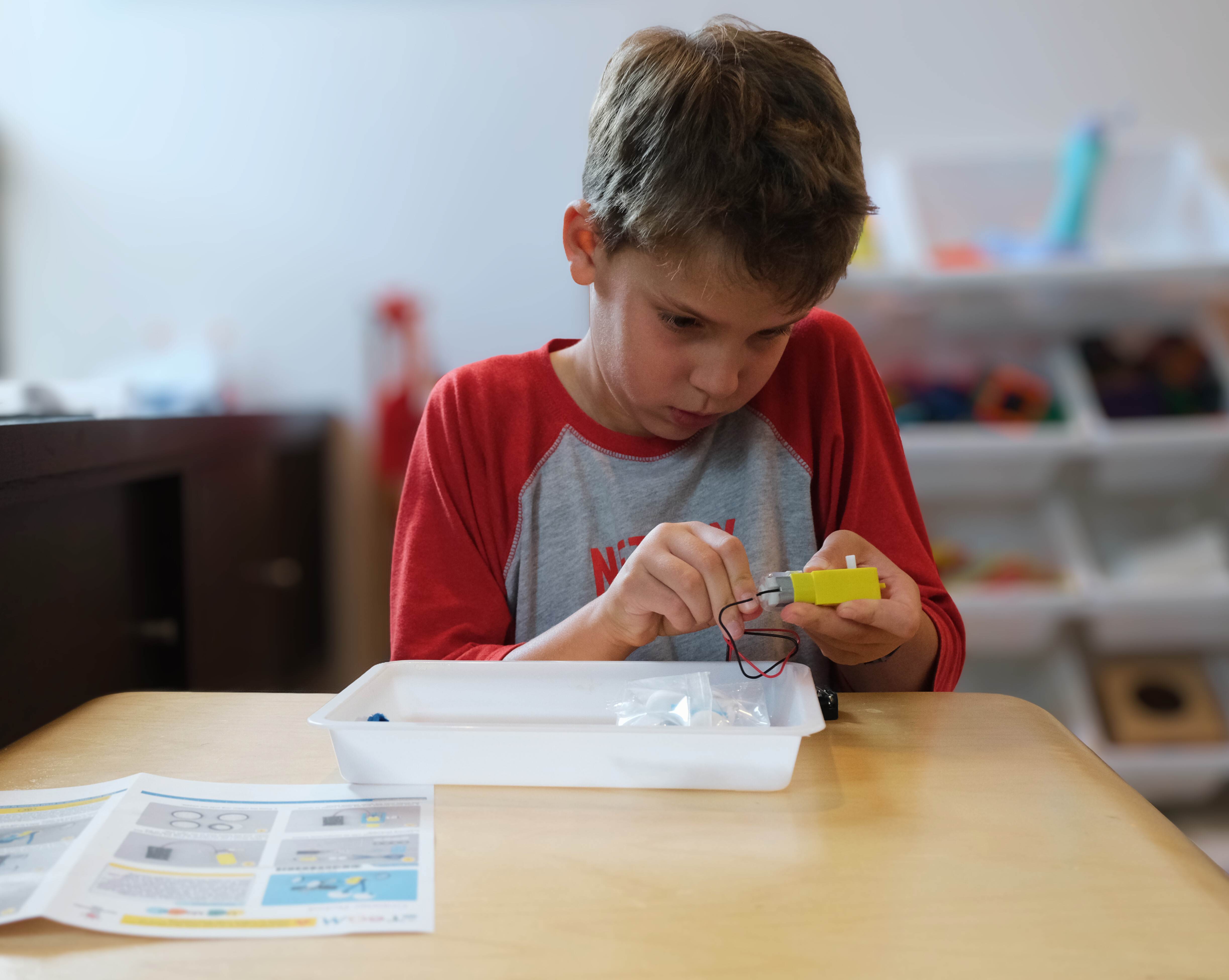 Boy in red and gray shirt assembling a yellow electronic device with wires at a table with instruction sheets.