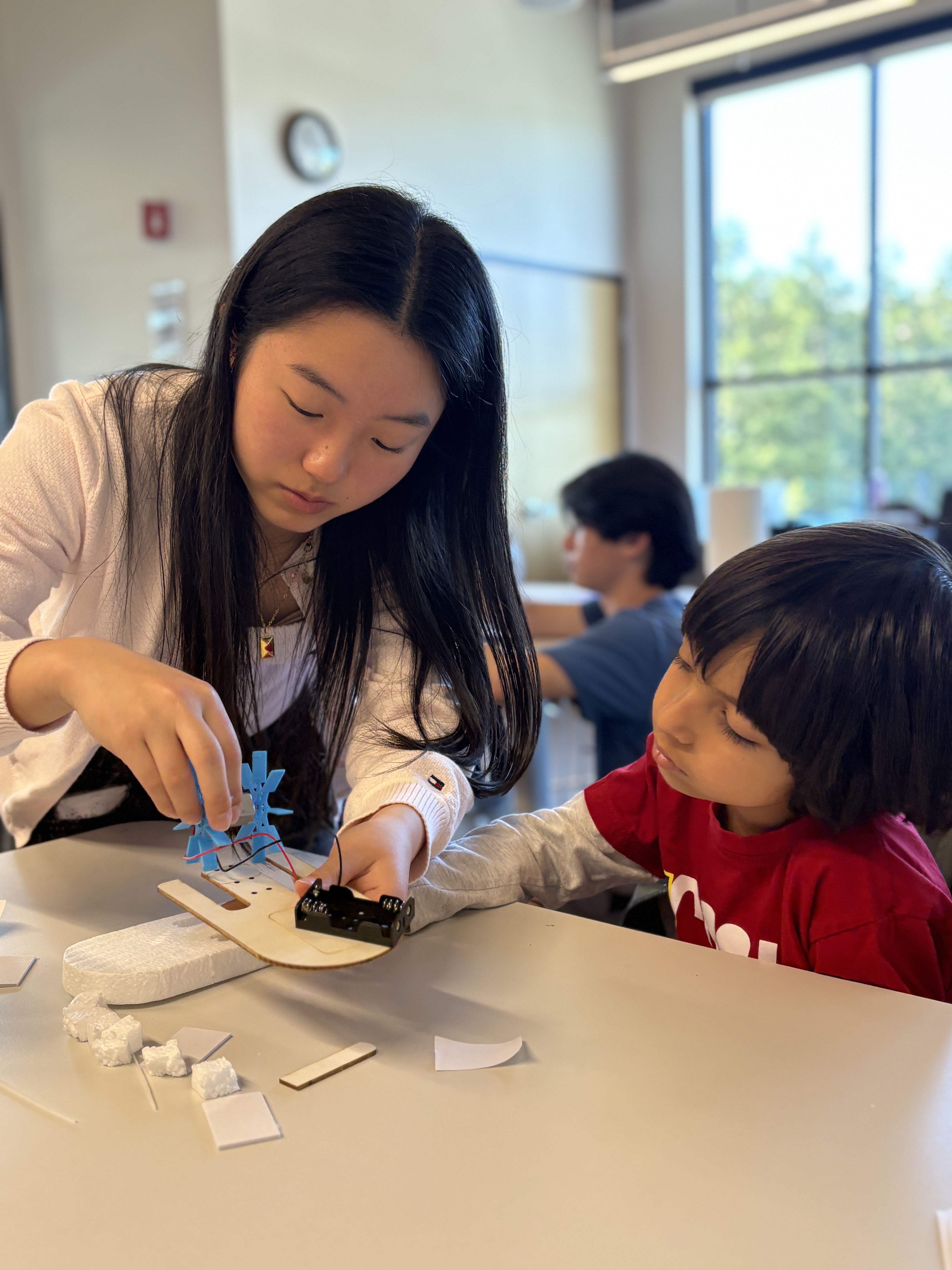 Two children working together on a small craft project with wires and blue plastic parts at a table indoors.