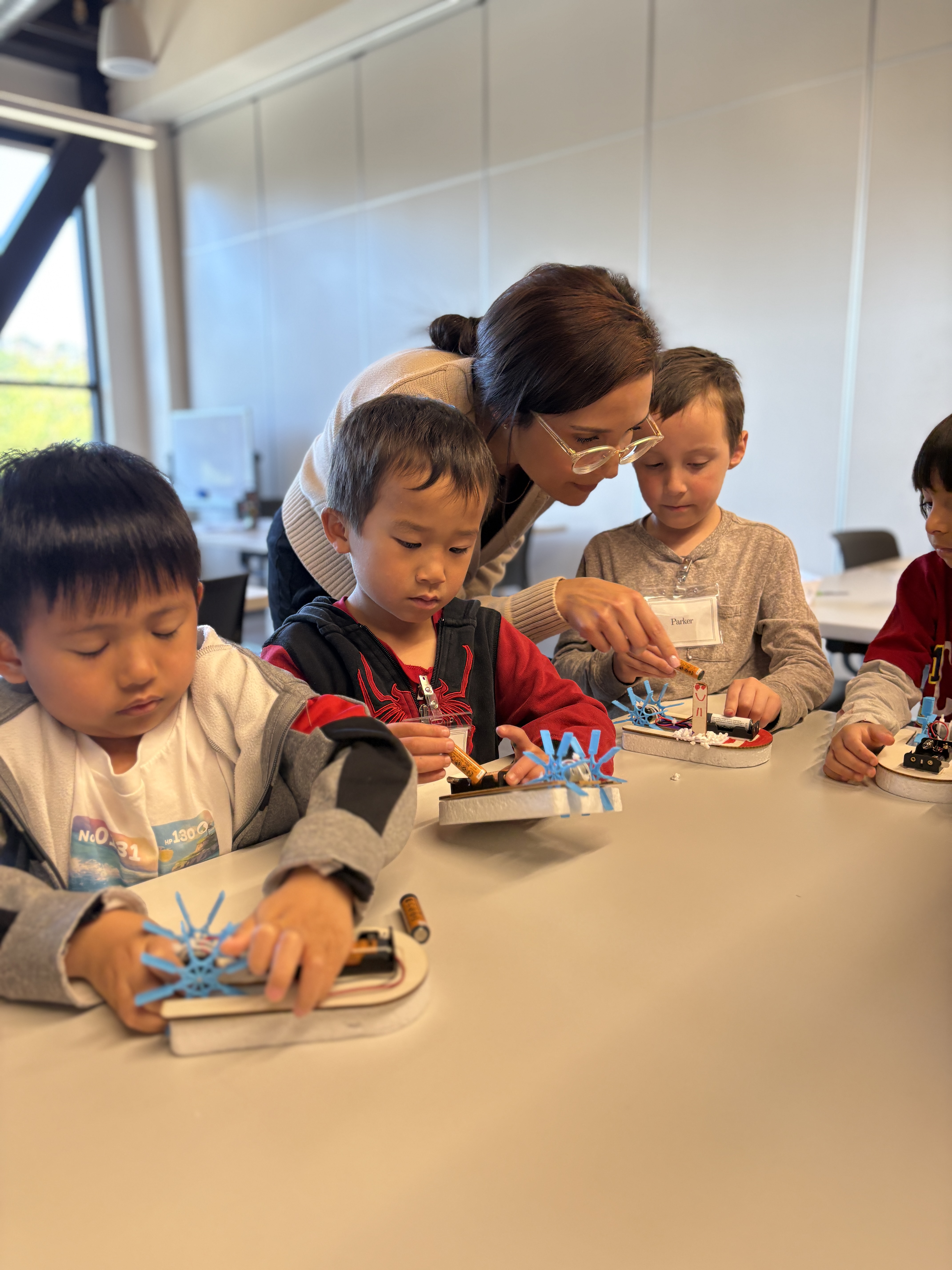 Teacher guiding three young boys working on a STEM project involving blue gears and batteries at a table.