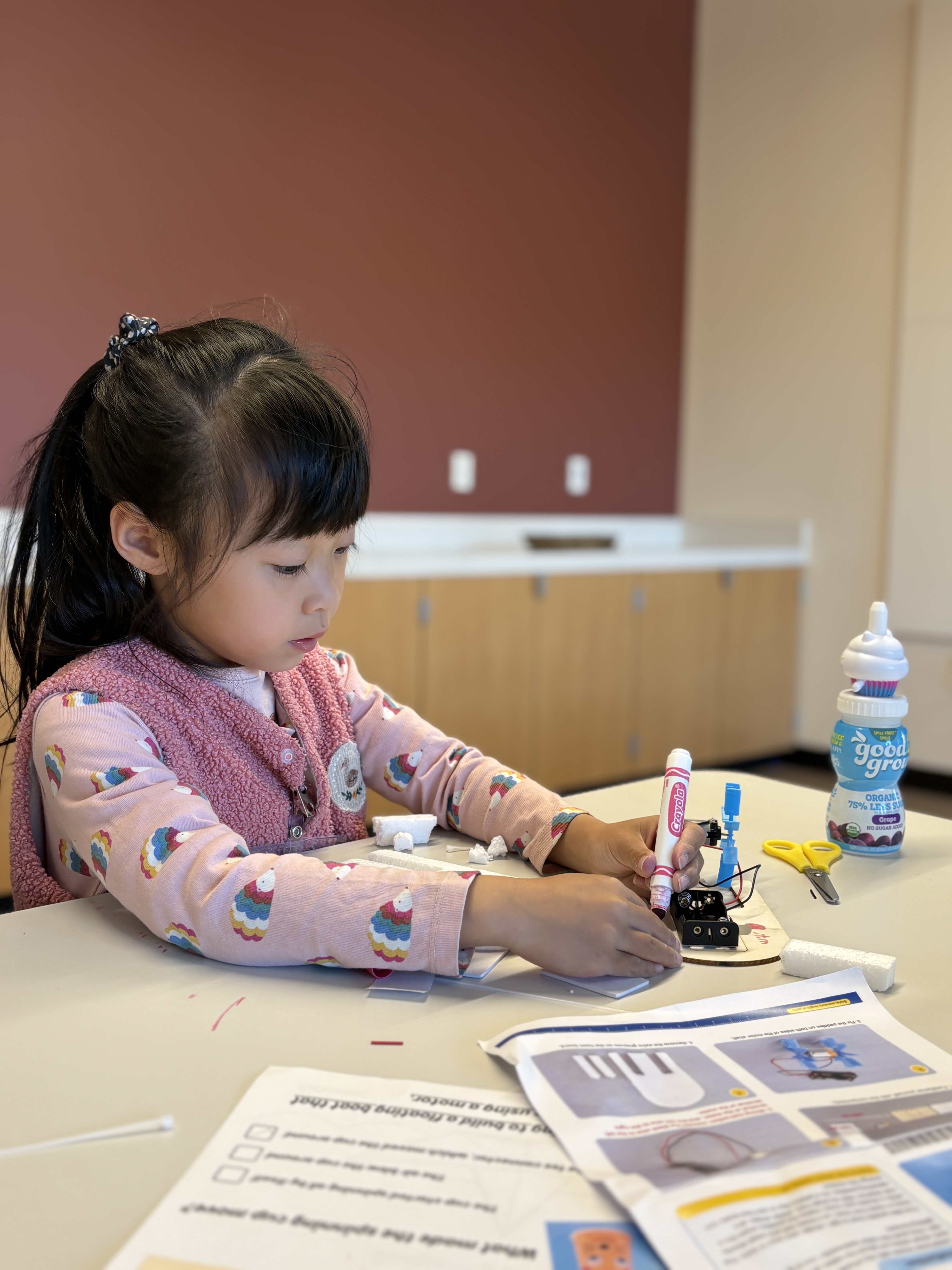 Young girl with a pink sweater and ponytail drawing on a small electronic circuit board at a table with instruction papers and craft supplies.
