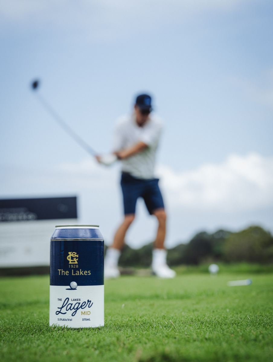 Can of The Lakes Lager Mid beer on grass with a golfer swinging a club blurred in the background.