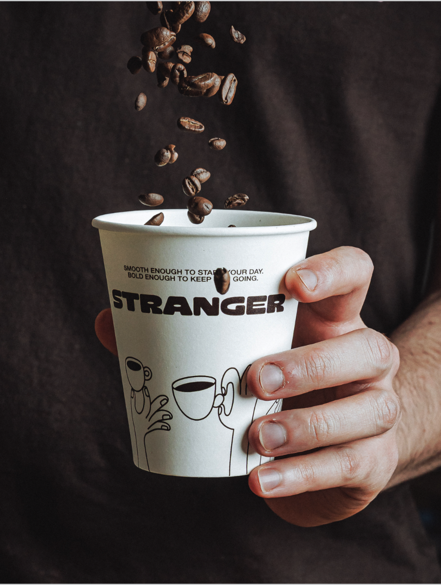 Hand holding a white coffee cup labeled 'STRANGER' with coffee beans falling into it.