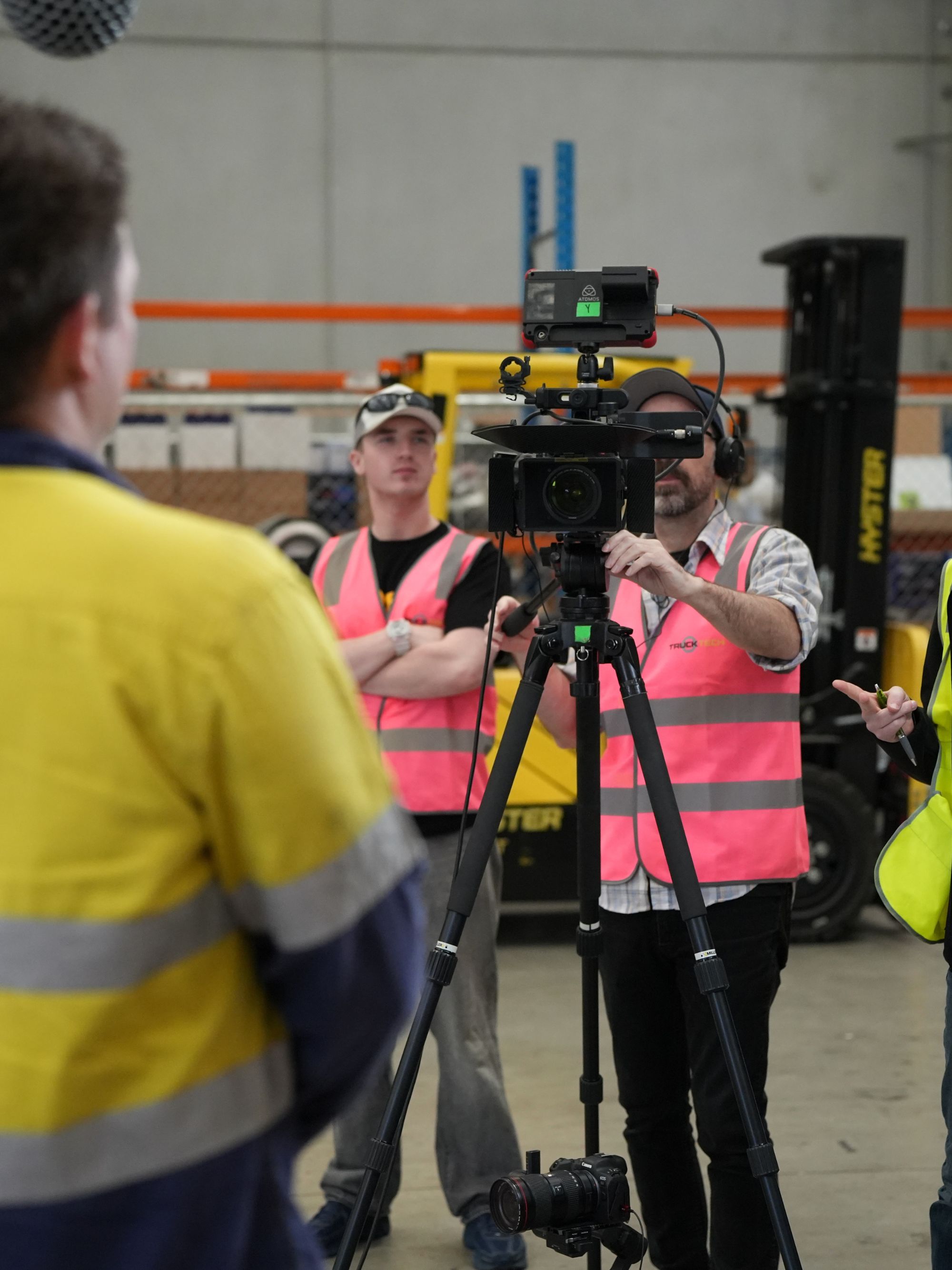 Man operating a camera on a tripod in a warehouse with others wearing safety vests.