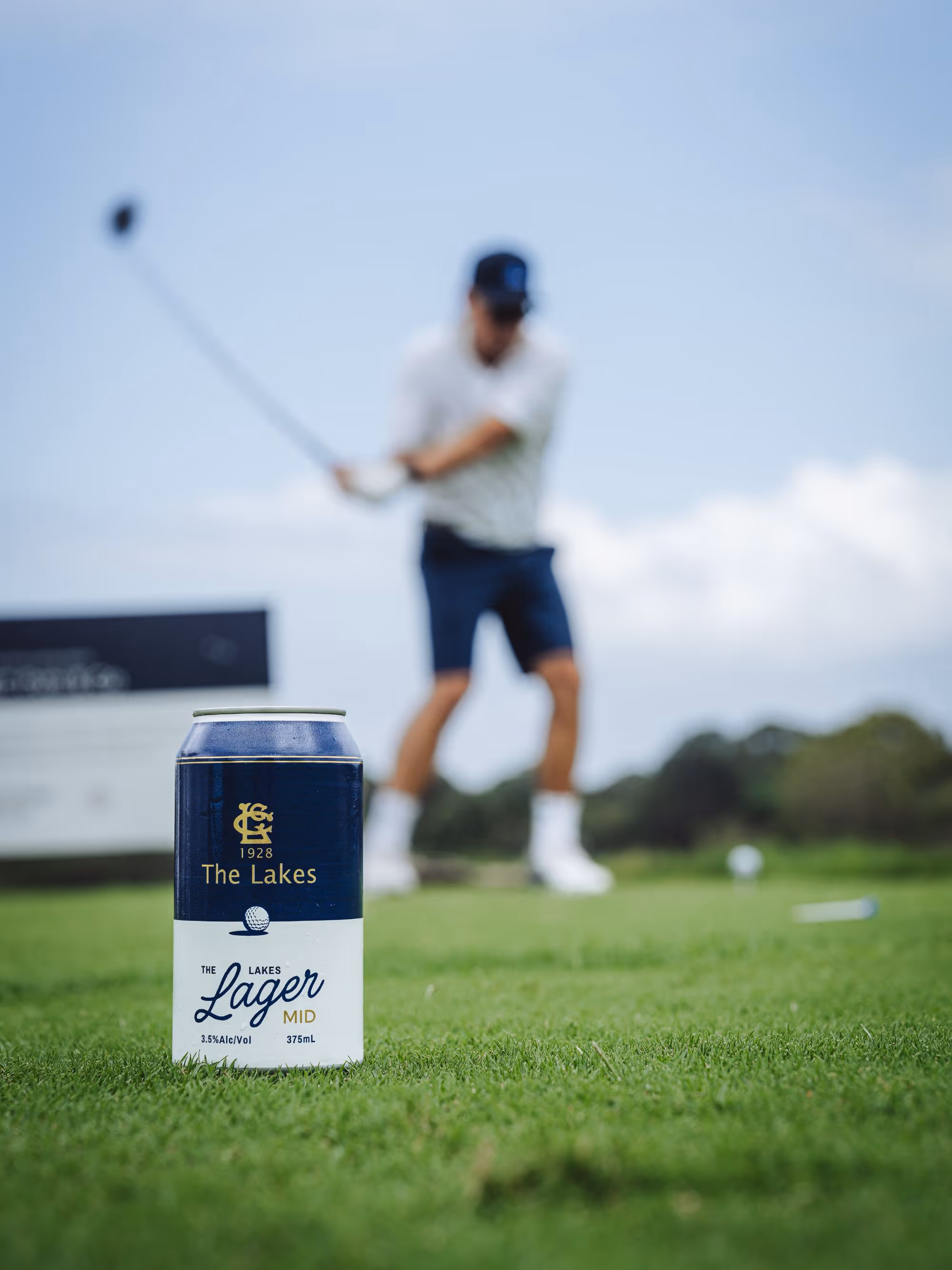 Can of The Lakes Lager Mid on grass with golfer swinging club in the background.