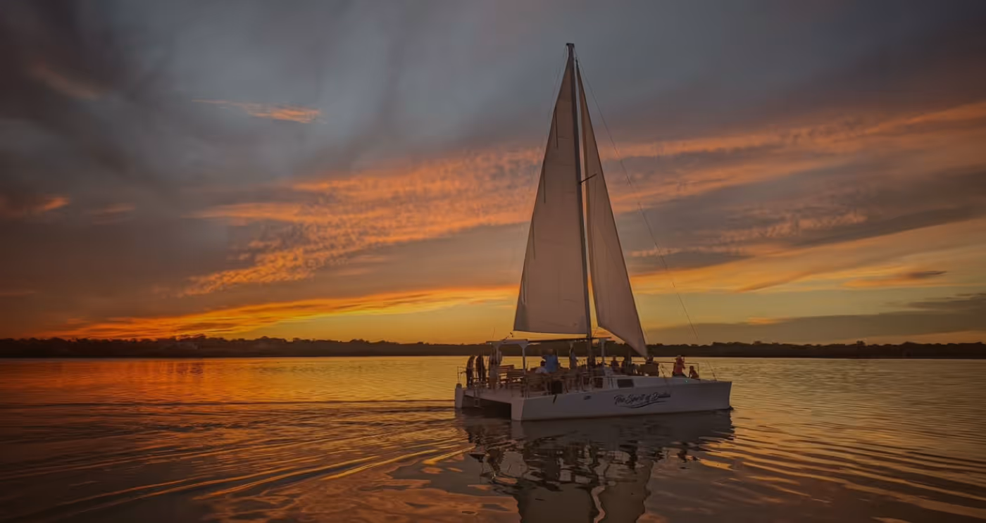 Sailboat with a large white sail on calm water during a colorful sunset with orange and purple skies.