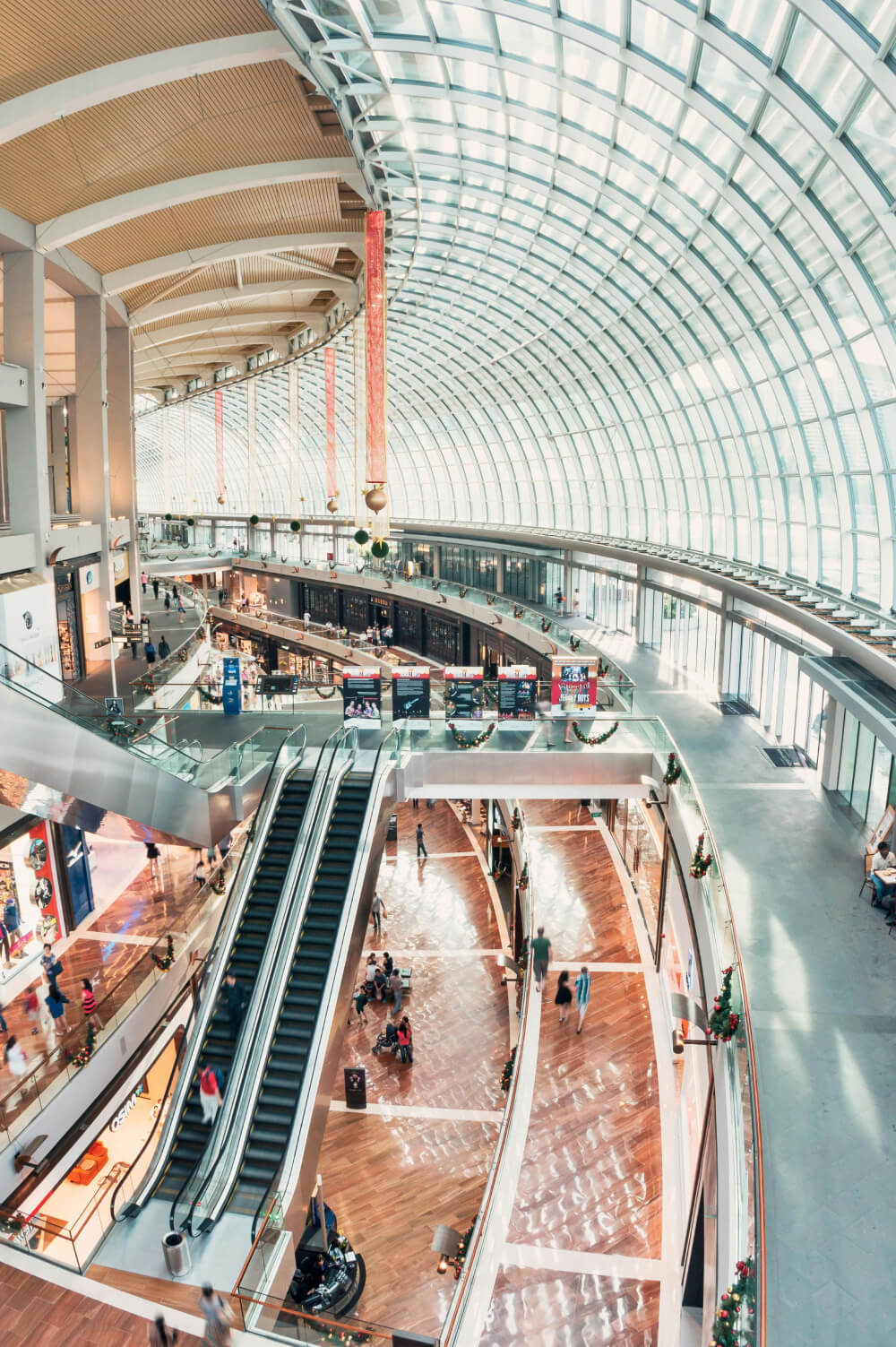 Interior view of a modern shopping mall with a large curved glass ceiling, escalators, and wooden flooring.