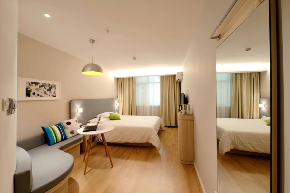 Modern hotel room with a double bed, round white table with laptop, curved sofa with colorful cushions, wooden floor, and large window with beige curtains.
