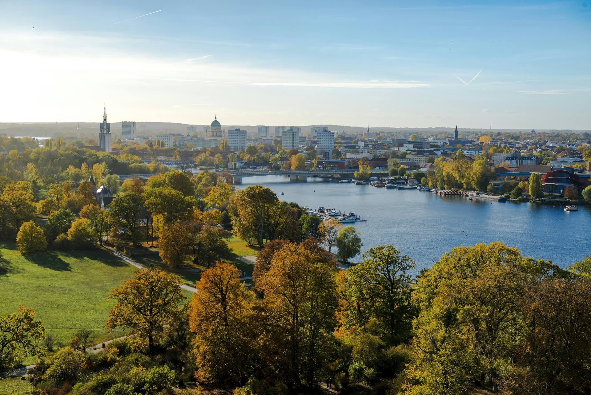 Panoramic view of a city with autumn trees, a river with boats, bridges, and buildings under a clear blue sky.