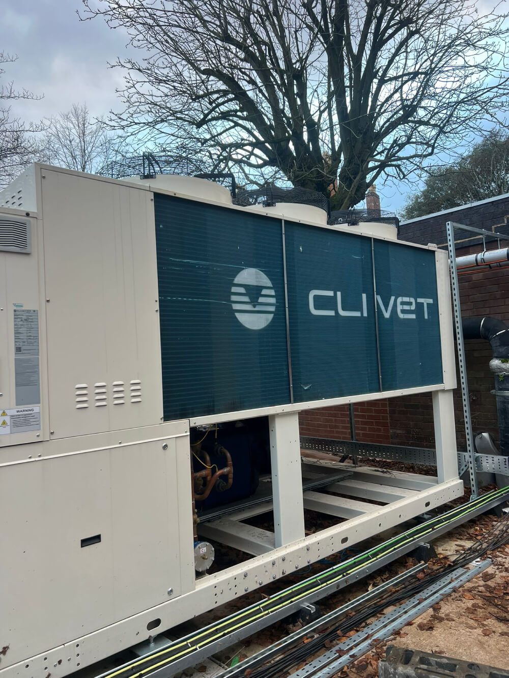 Large outdoor Clivet HVAC unit with visible fans on top and pipes underneath next to a brick wall and leaf-covered ground.