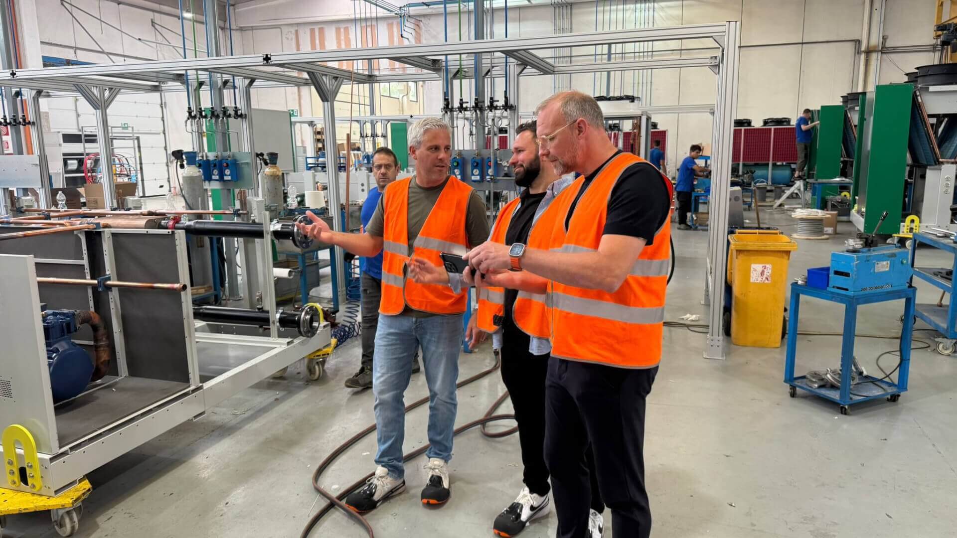 Three men wearing orange safety vests discussing inside an industrial workshop with machinery and equipment around.