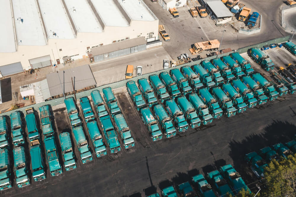 Aerial view of numerous green garbage trucks parked in organized rows next to a large warehouse building.