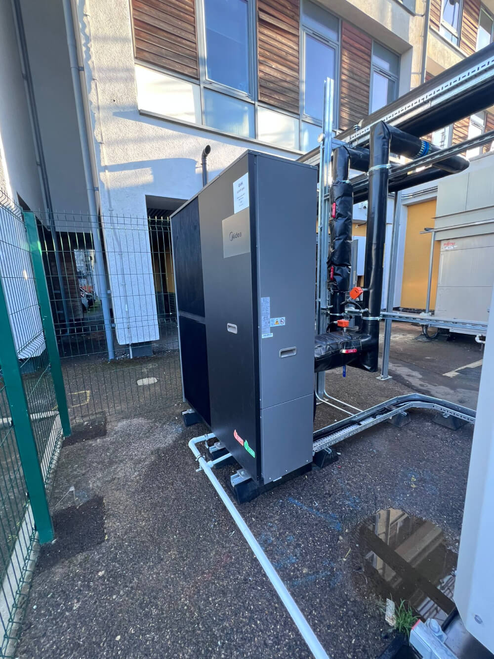 Outdoor industrial HVAC unit connected to insulated pipes near a building with windows.