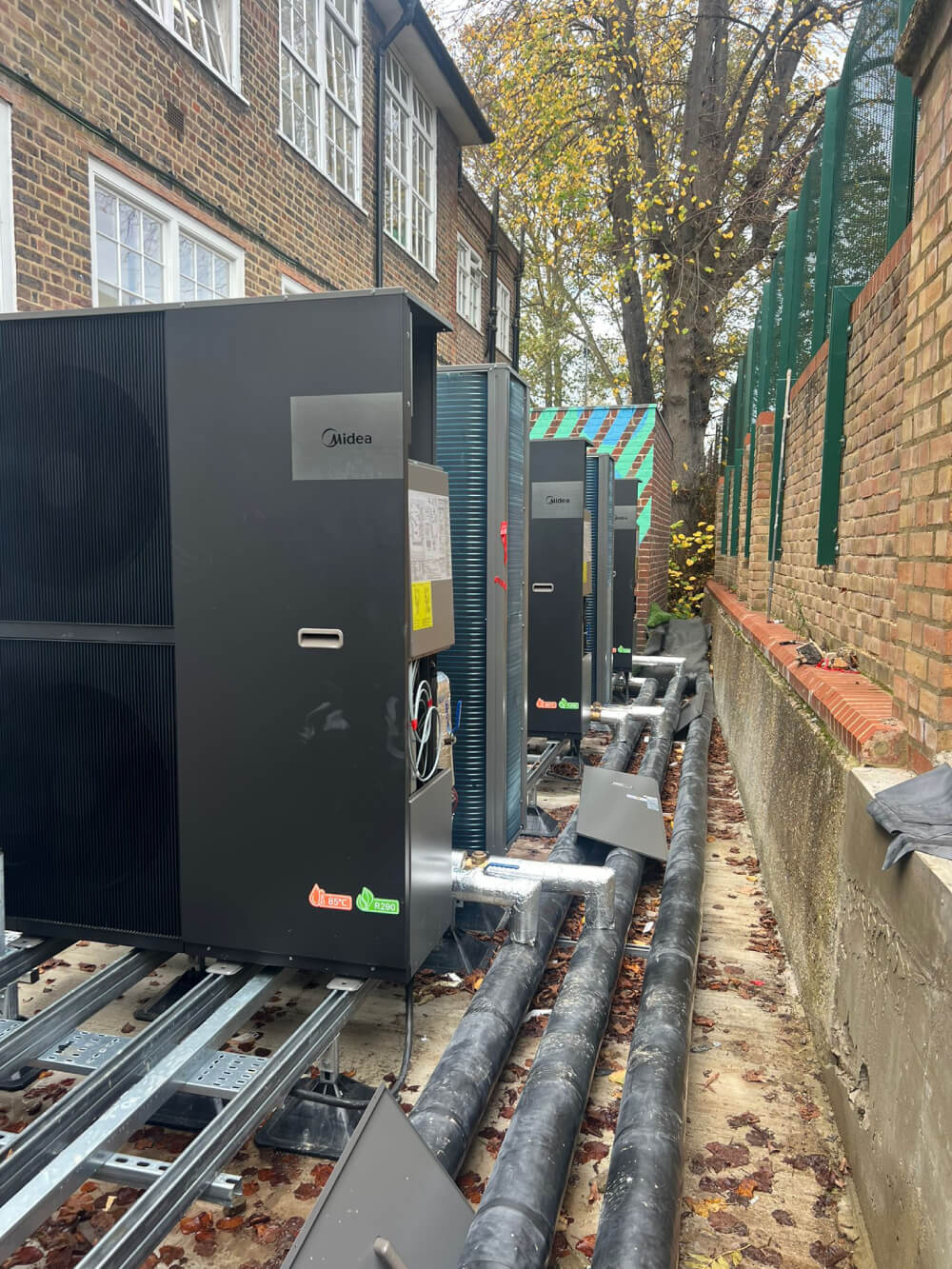 Multiple black Midea HVAC units installed outdoors beside a brick building and a fence, with insulated pipes running along the ground.