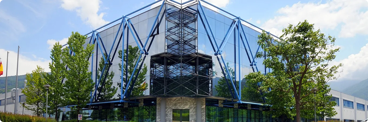Modern glass building with blue structural supports surrounded by green trees under a partly cloudy sky.