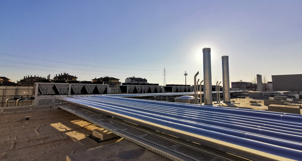 Industrial rooftop with metal pipes, ventilation units, and chimneys under a clear blue sky with the sun shining.