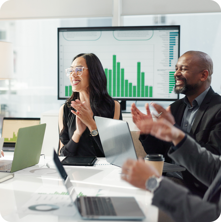 Business team clapping and celebrating success during a meeting with laptops and a screen showing green bar graphs.