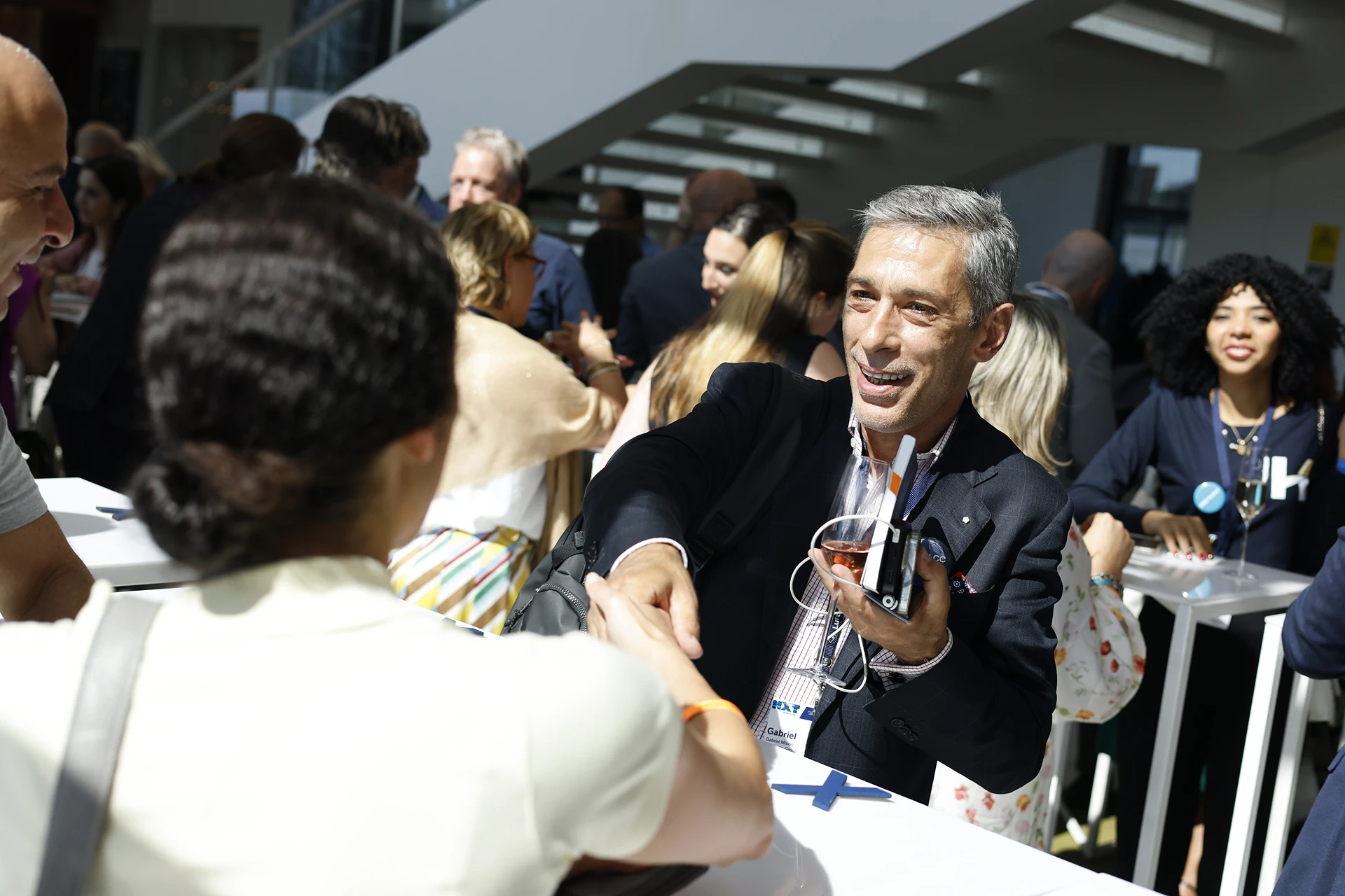 Man in a suit smiling and shaking hands with a woman at a busy social event with people conversing in the background.