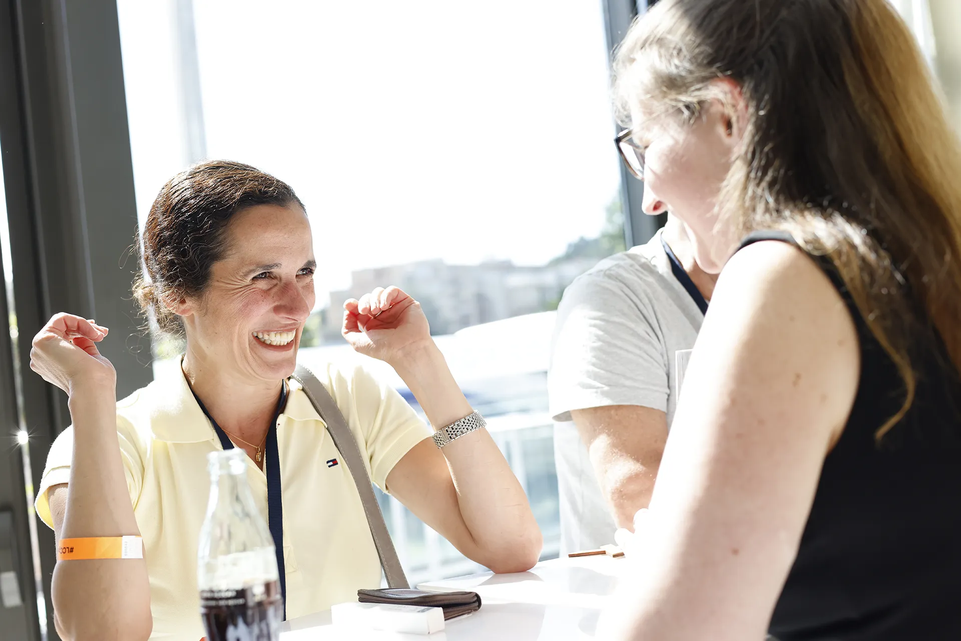 Two women smiling and engaging in conversation at a sunlit table with a glass bottle and a wallet.