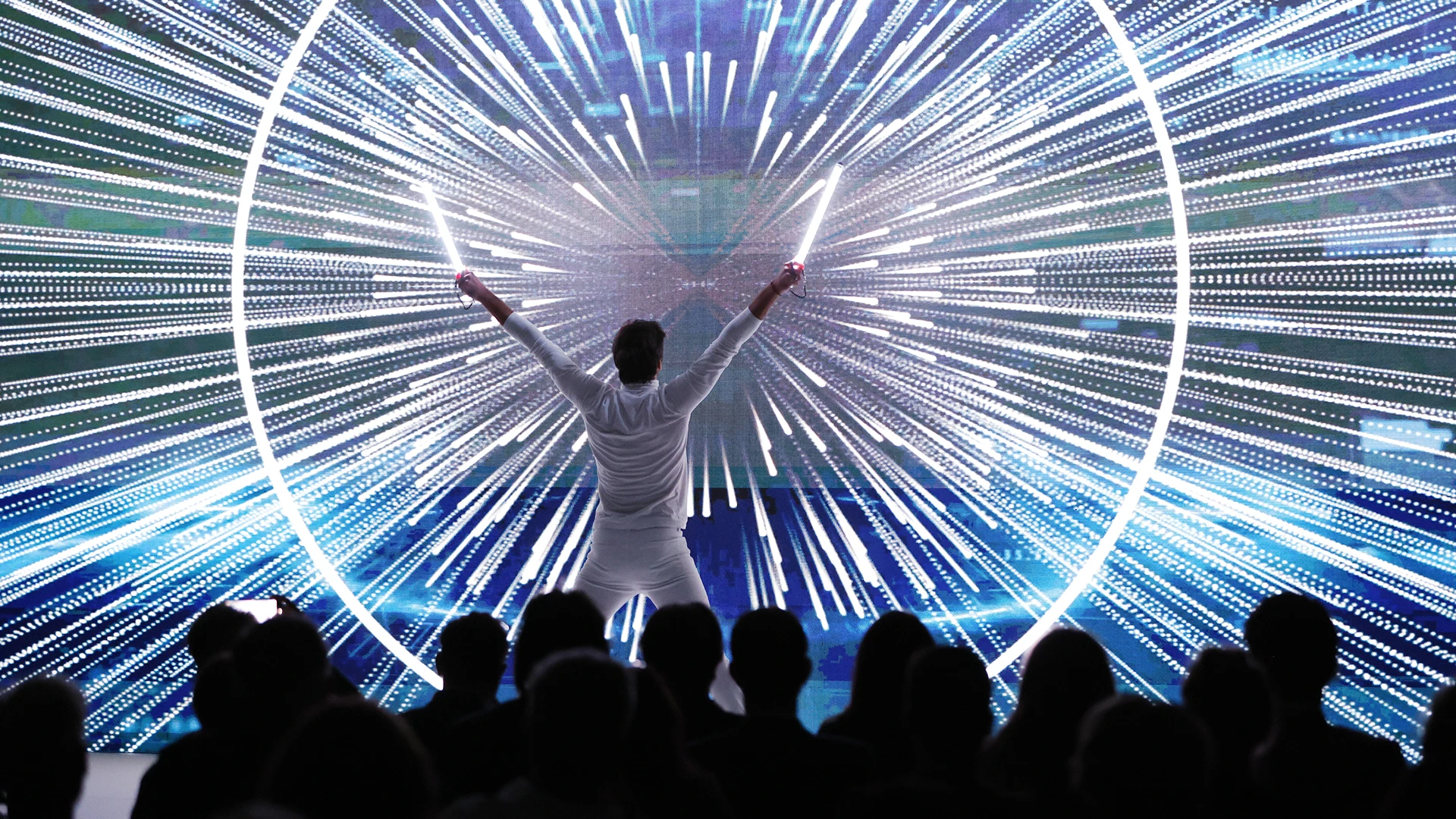 Person in white outfit holding two light sticks raised with an audience watching and radial light beams displayed on a large screen behind.
